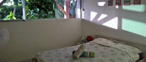 A bedroom with a bed, a fan, a window, and a wall with framed pictures.