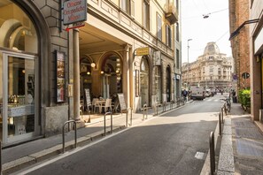 Uma rua com um edifício com entrada em arco, um letreiro de 'Officine Ortopediche Milano' e vista de um grande edifício ao fundo.