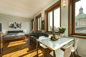 Une chambre d’hôtel moderne équipée d’un lit, d’un canapé, d’une table à manger, de chaises et de grandes fenêtres donnant sur la ville.