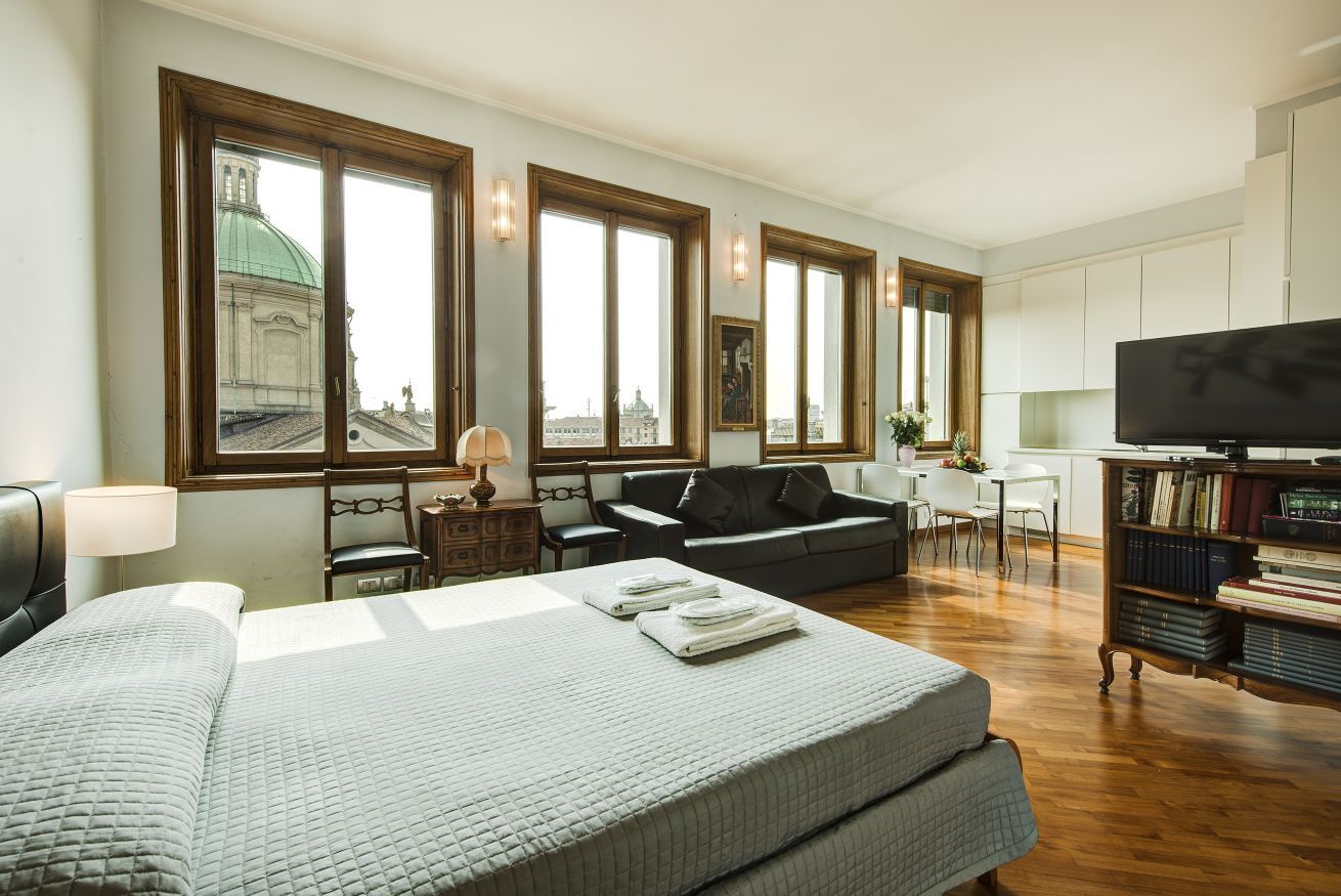 A hotel room with a large bed, a sofa, a TV, a bookshelf, and large windows with a view of a building.