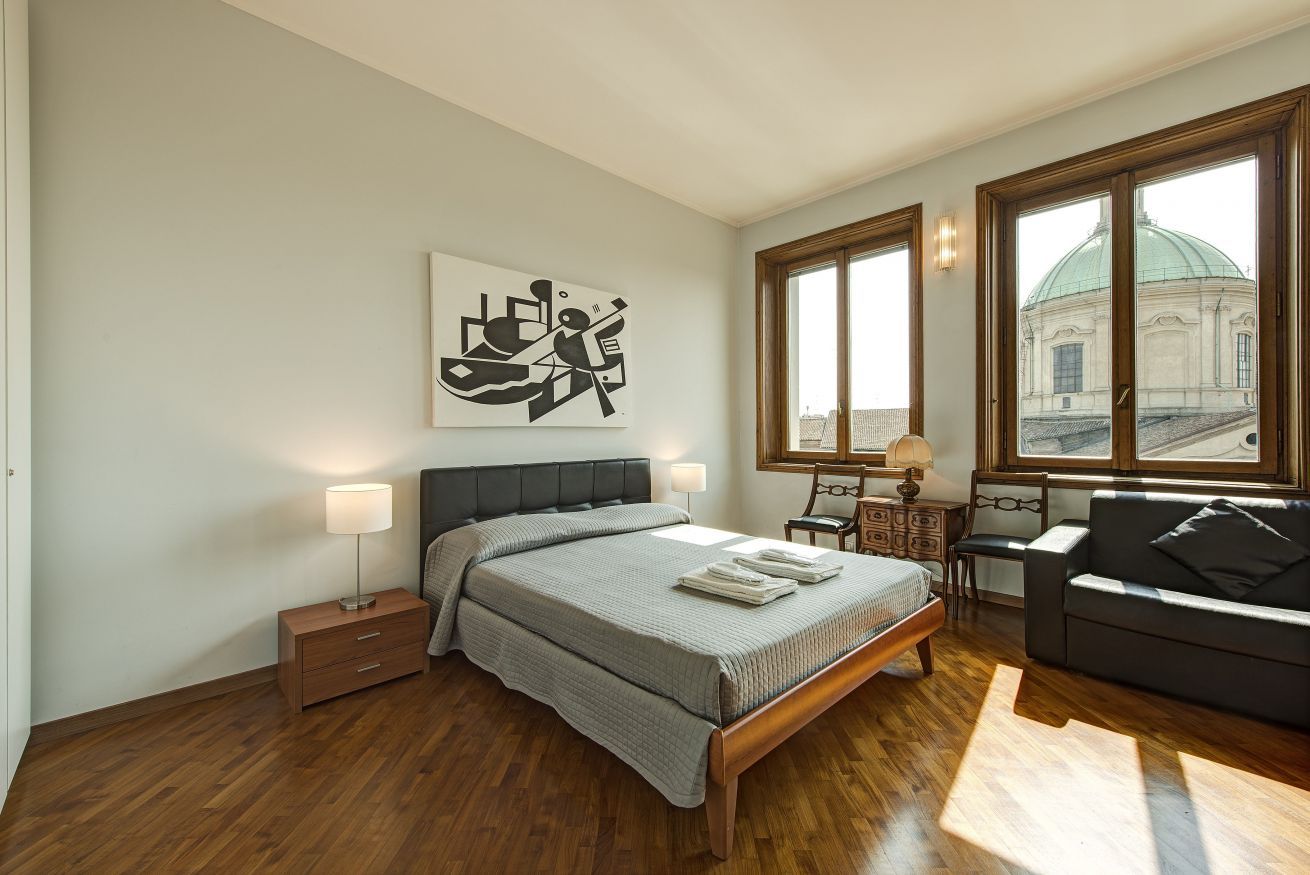 Un dormitorio con cama, mesitas de noche, sofá, ventana con vista a un edificio y una obra de arte en la pared.