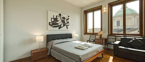 Un dormitorio con cama, mesitas de noche, sofá, ventana con vista a un edificio y una obra de arte en la pared.