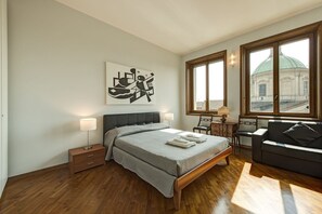 Een slaapkamer met een bed, nachtkastjes, een bank, een raam met uitzicht op een gebouw en een kunstwerk aan de muur.