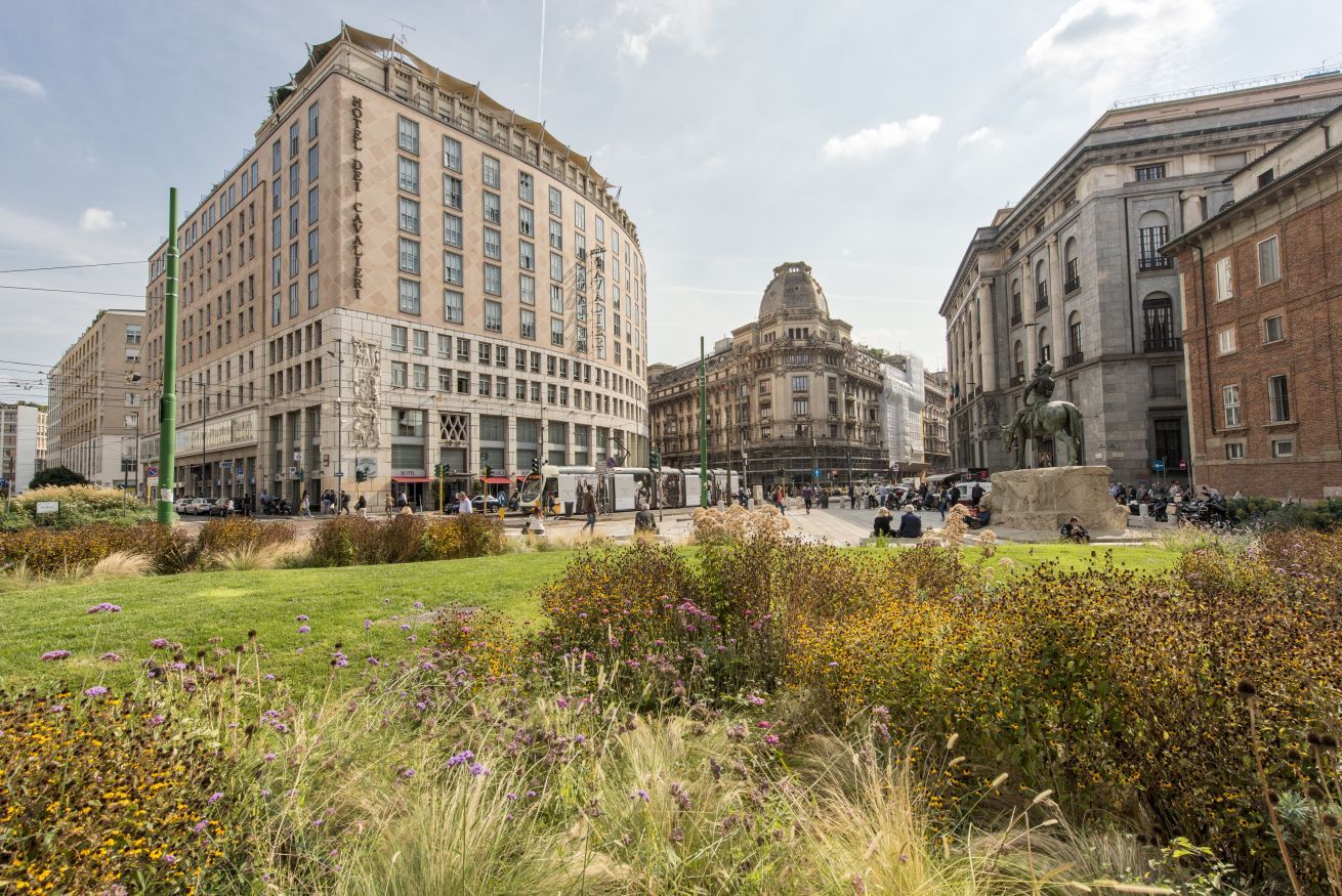 Ein historisches Gebäude mit einer zentralen Kuppel, umgeben von einem Platz mit einer Statue und einem Brunnen.