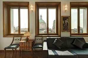 A room with a wooden desk, two chairs, a sofa, and three large windows with views of a cityscape and a building with a dome. - Panoramic apartment equipped with every comfort 2 minutes from the Duomo (Milano)