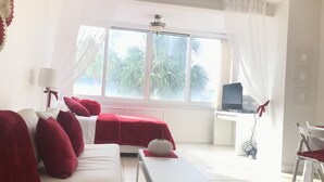 Una camera d'albergo con un letto, una scrivania con un computer e una sedia. Si vede una finestra con tende e, all'esterno, una vista su palme.