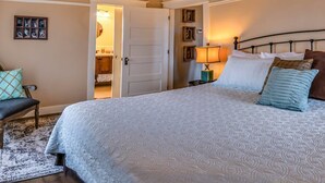 Ein Schlafzimmer mit einem großen Bett, einem Sessel, einer Tür zum Badezimmer und einem kleinen Tisch mit Lampe.