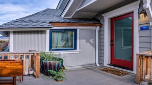 Ein Haus mit roter Tür, einem Fenster mit blauer Zierleiste und einer kleinen Veranda mit Holztisch und Topfpflanzen.