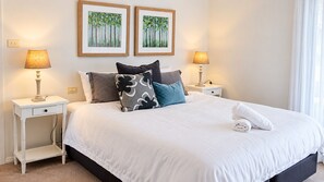 A neatly made bed with white linens, two bedside tables with lamps, and two framed paintings on the wall. - Beachside Haven - Your Perfect Oasis Home (Corlette)