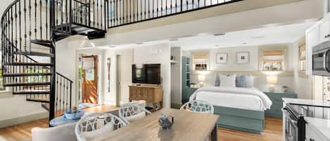 A spacious living area with a wooden dining table, white wicker chairs, a large bed with a blue headboard, and a black spiral staircase.
