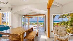 Una habitación amplia con una mesa de madera, bancos y una silla de mimbre. Cuenta con ventanas grandes que ofrecen vista a la playa y al océano.