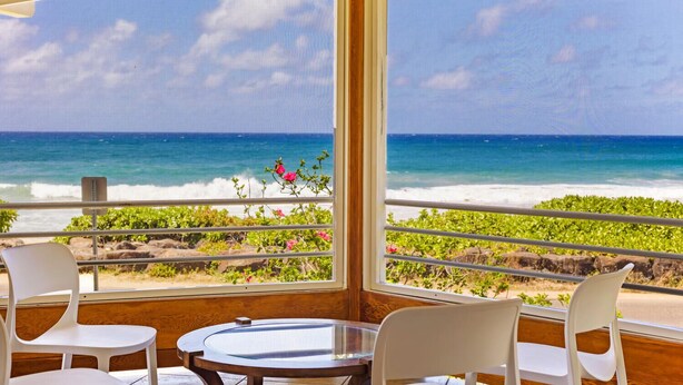 Una habitación con un ventanal grande que da a la playa, dos sillas blancas y una mesita.