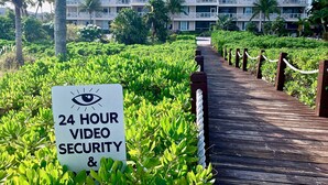 Un panneau indiquant la présence d’un système de vidéosurveillance en service 24 heures sur 24, ainsi qu’un chemin menant à un bâtiment doté de balcons.