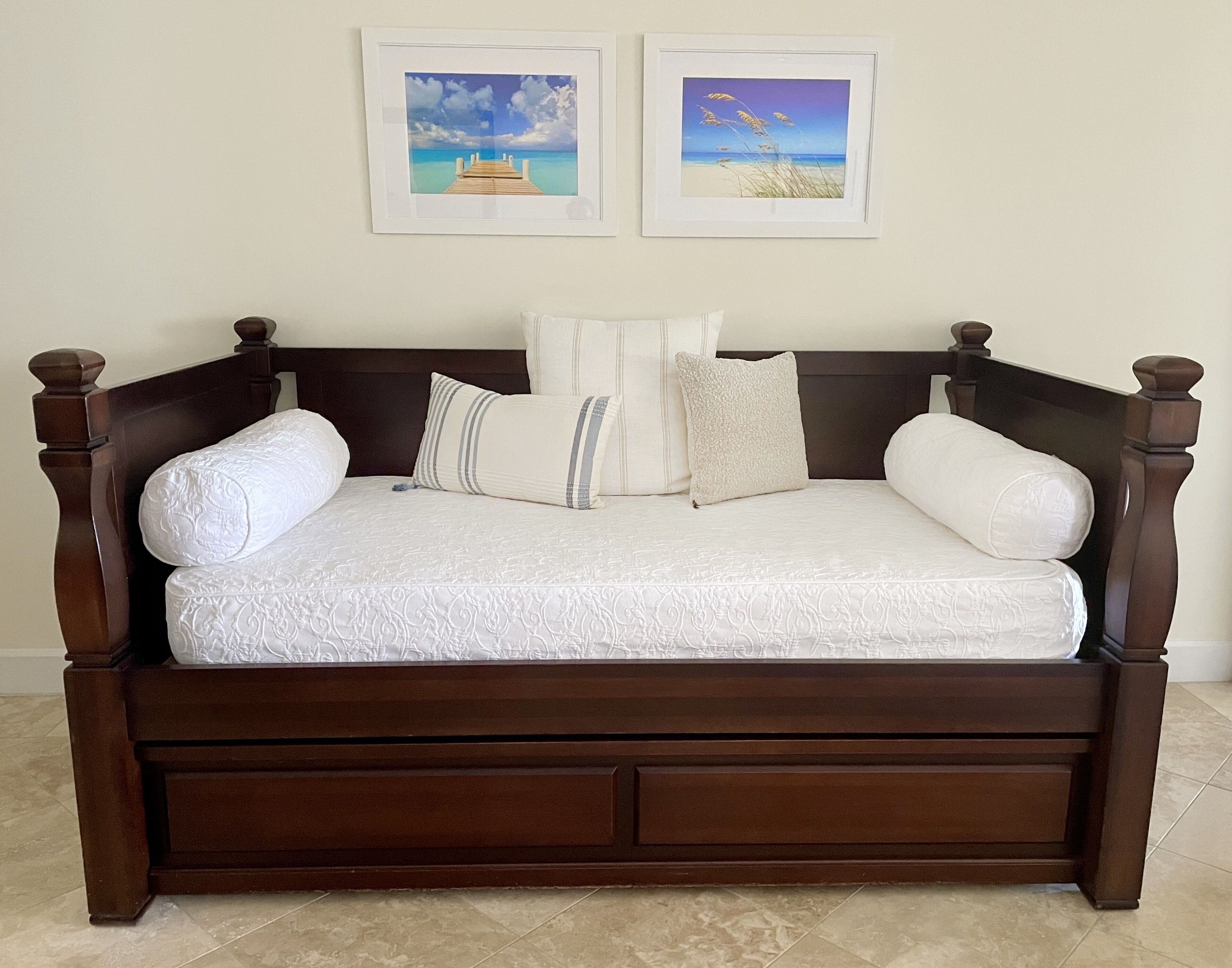 A dark wood four-poster bed with a white quilt and pillows, two framed pictures on the wall above, and a tiled floor.