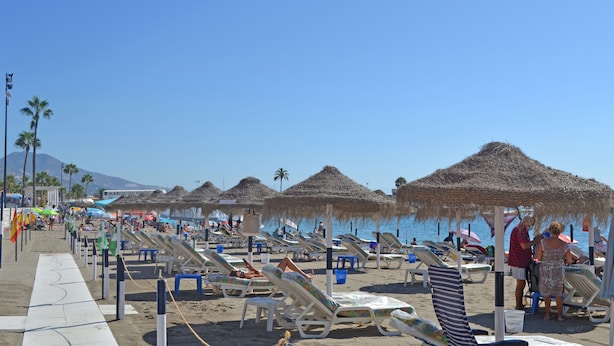 Ein Strand mit Liegestühlen und Reetdachsonnenhütten, bewölkter Himmel und Palmen.