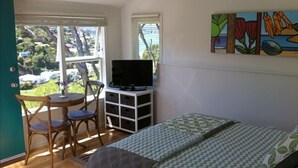 A room with a bed, a TV on a white cabinet, a dining table with chairs, and a window with a view of trees and houses.