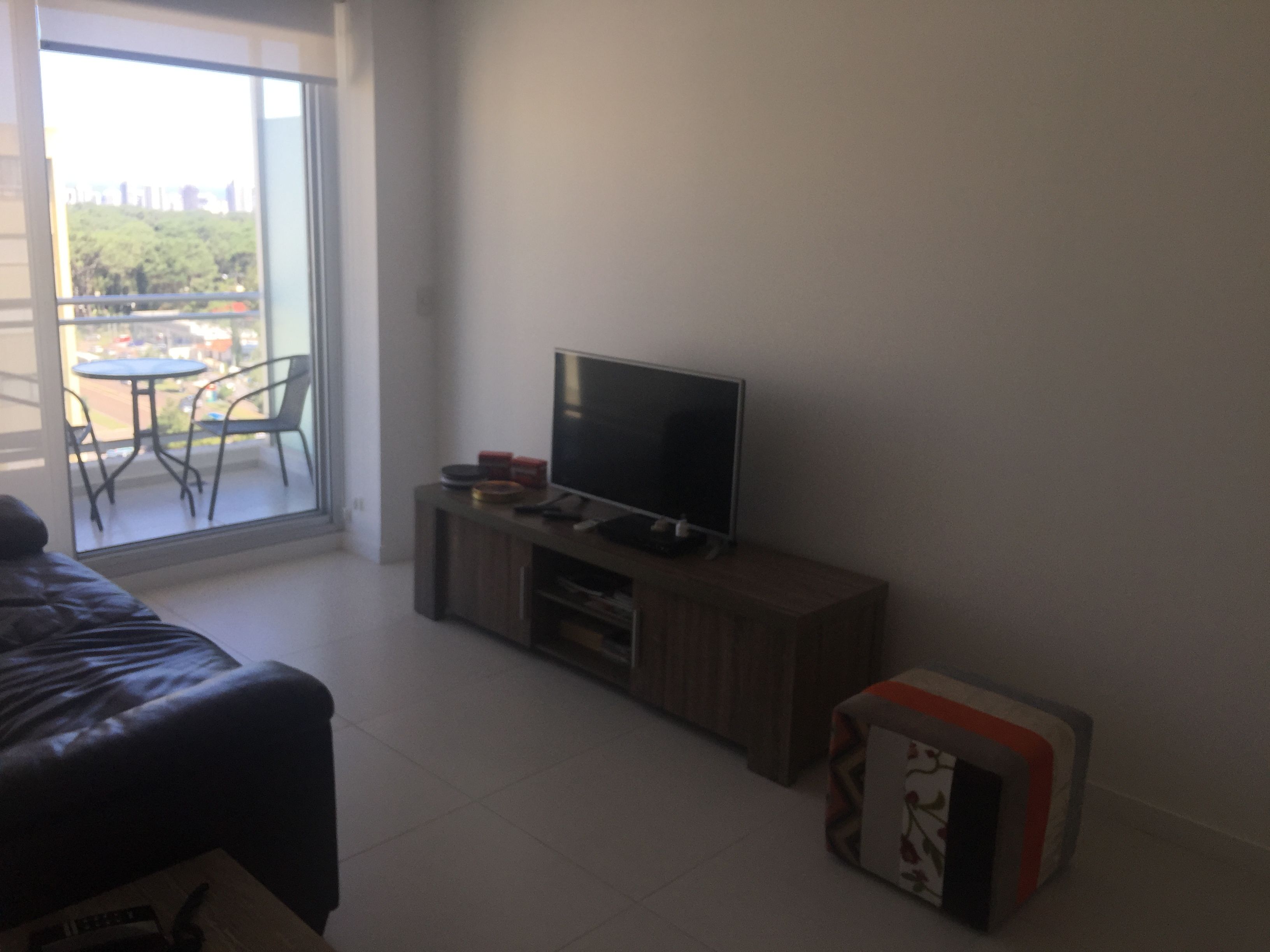 Habitació amb un llit, una televisió sobre un suport de fusta, una taula petita amb cadires i un balcó amb vista a arbres i edificis.