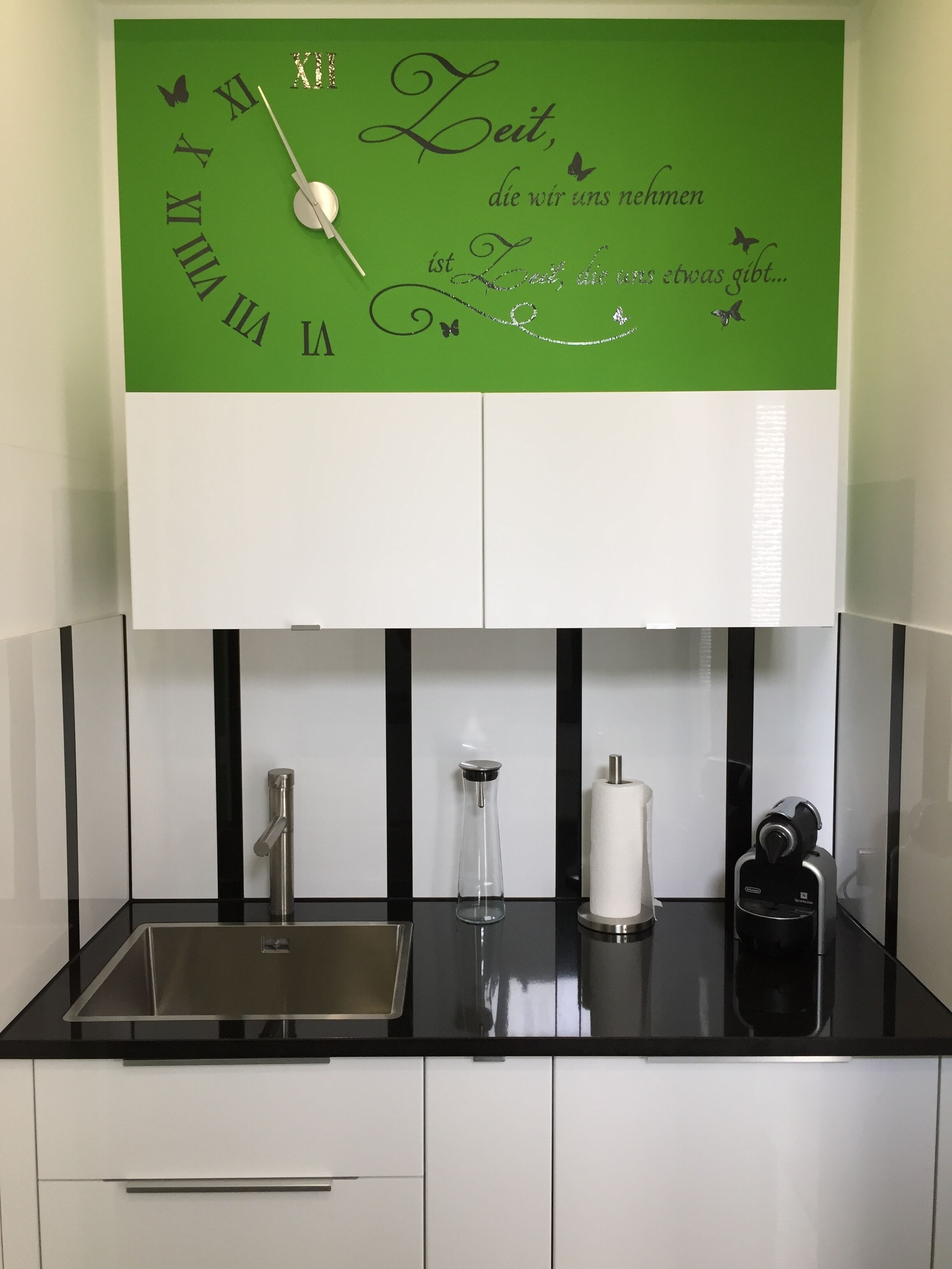 A kitchen with a green wall clock, a stainless steel sink, a glass vase, and a coffee machine.