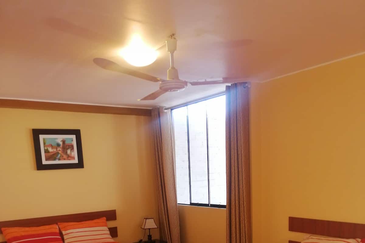 Quarto de hotel com duas camas, um ventilador de teto, uma janela com cortinas e um quadro na parede.