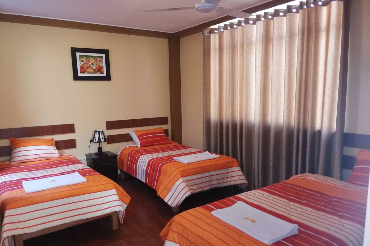 Quarto de hotel com duas camas, cada uma com roupa de cama listrada, uma mesa de cabeceira com abajur, um ventilador de teto e um quadro na parede.