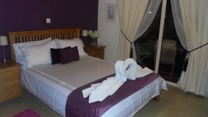 A neatly made bed with white linens, a purple throw blanket, and two white towels arranged in a heart shape. There is a wooden nightstand with a lamp and a small vase with flowers beside the bed. - Luxury 5* apartment overlooking 2 swimming pools.  (Peyia)