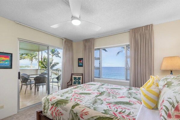 A hotel room with a bed, a balcony with chairs, a view of the sea, and a ceiling fan. - Revel in Unparalleled Views of Maui's Coastal Majesty! - Royal Mauian 416 (Kihei, Maui)
