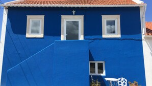 Un edificio azul con techo de tejas rojas, ventanas de marco blanco y un pequeño espacio al aire libre con sillas y mesas de metal blanco.