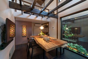 Un espace de salle à manger moderne avec une table en bois, des chaises et des luminaires suspendus. On aper çoit un escalier à l’arrière-plan, qui mène à un niveau supérieur.