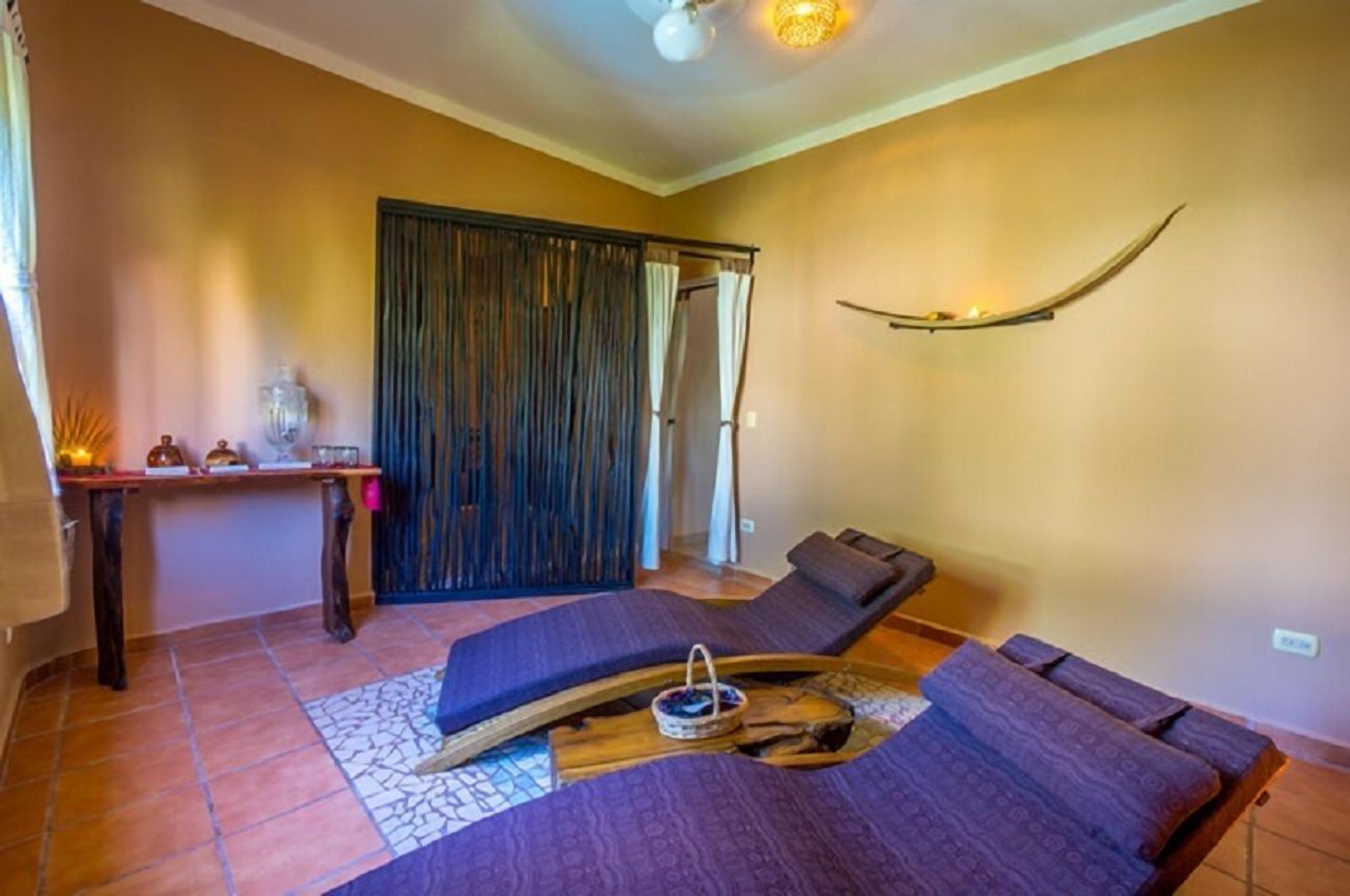 A room with two reclining beds, a wooden table with a tray, a decorative wall piece, and a curtain-covered door.