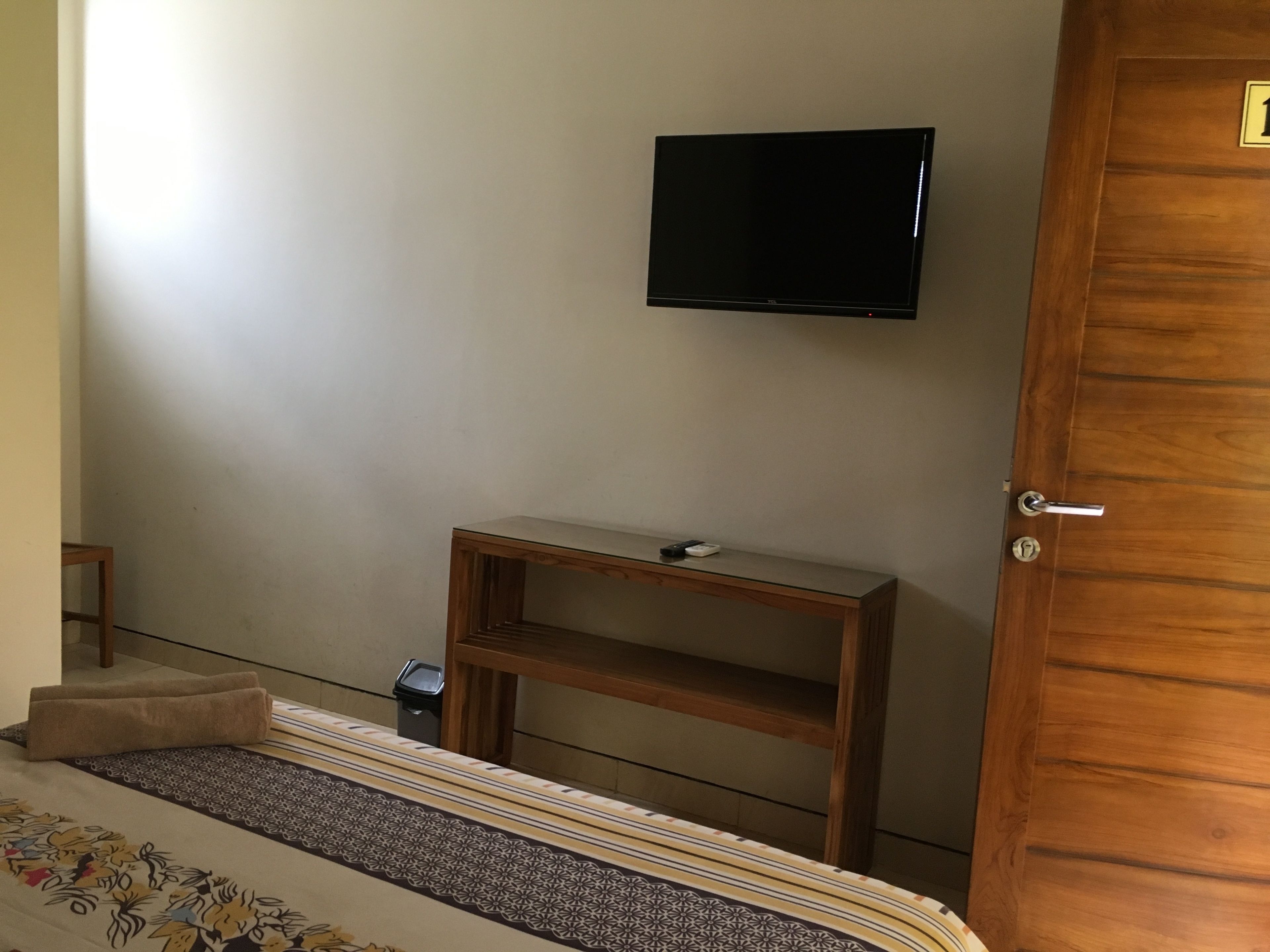Un dormitorio con una cama, una mesita de noche de madera, un televisor montado en la pared y una puerta.