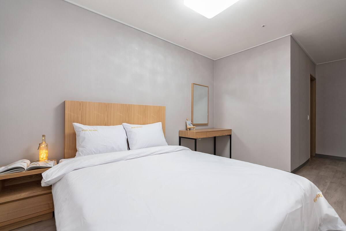 A modern hotel room with a large bed, a wooden nightstand with a book and a bottle, a small table with a mirror, and a light fixture.