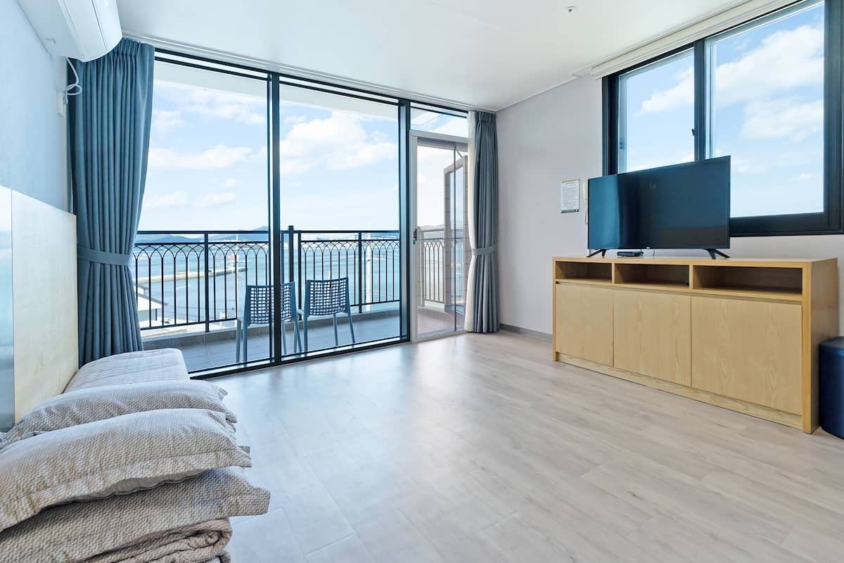 A spacious room with a large sliding glass door leading to a balcony with a view of the sea. A flat-screen TV is mounted on the wall, and there is a wooden cabinet with a TV stand.