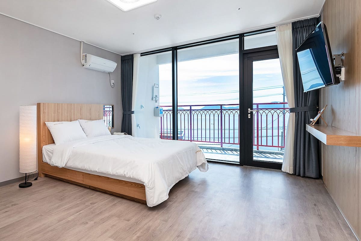 A modern hotel room with a wooden bed, a large window with a balcony, and a flat-screen TV mounted on the wall.