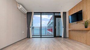 A modern hotel room with a wooden floor, a flat-screen TV mounted on a wooden panel, and a large sliding glass door leading to a balcony with a red railing. - Gold Beach Resort (Yeosu)