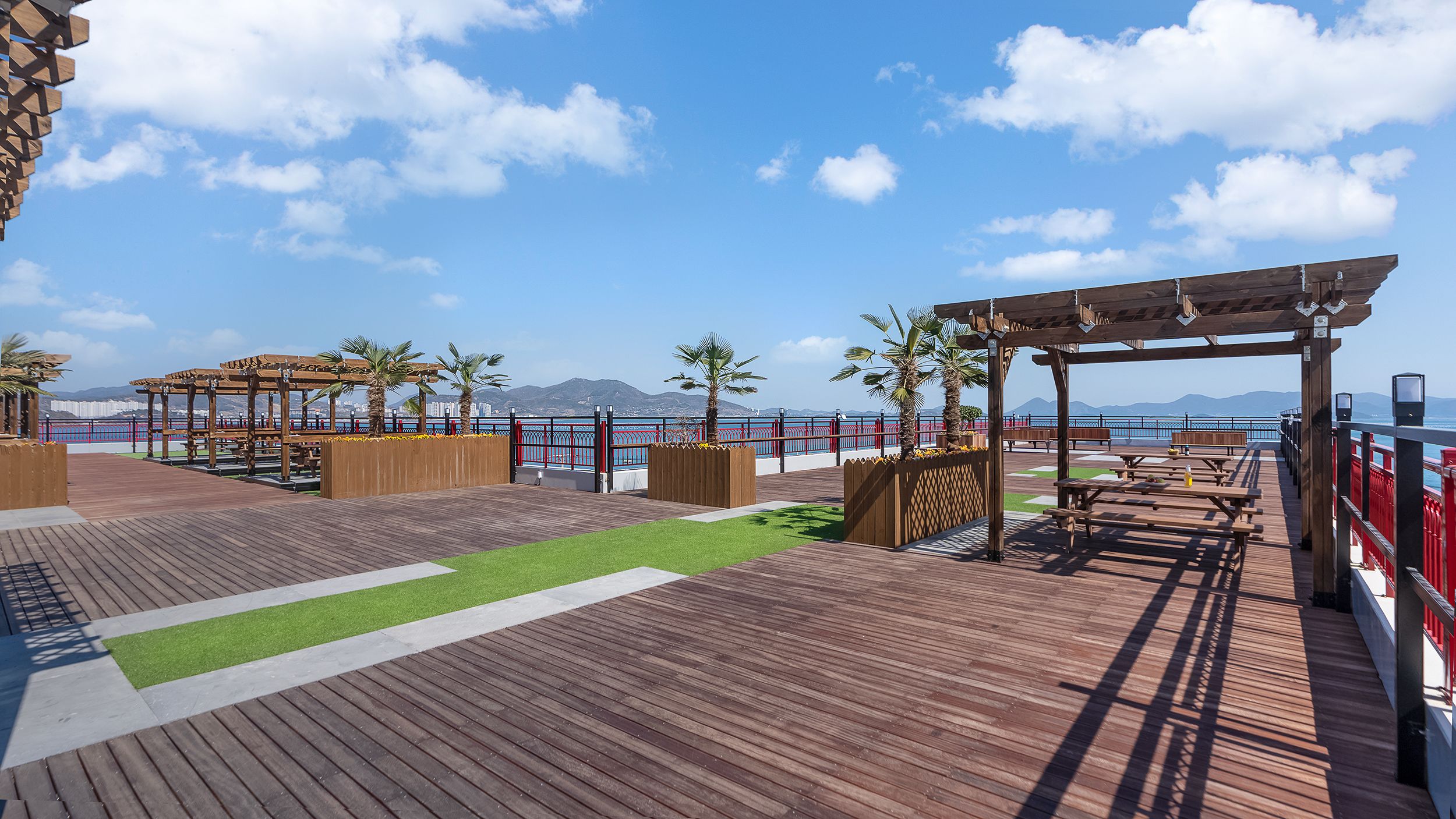 Une terrasse en bois avec des tables de pique-nique et des bancs, des palmiers et une vue sur la mer.