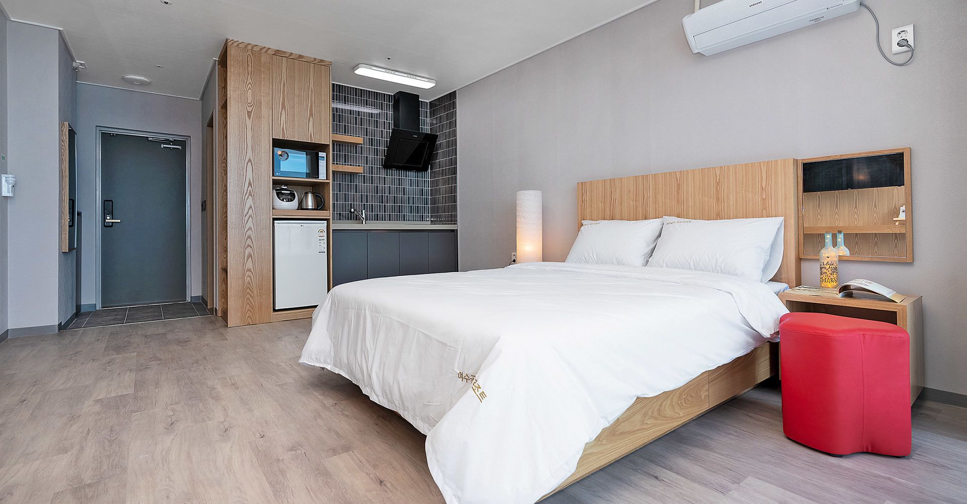 A modern hotel room with a large bed, a wooden wardrobe, a small kitchenette, and a red ottoman.
