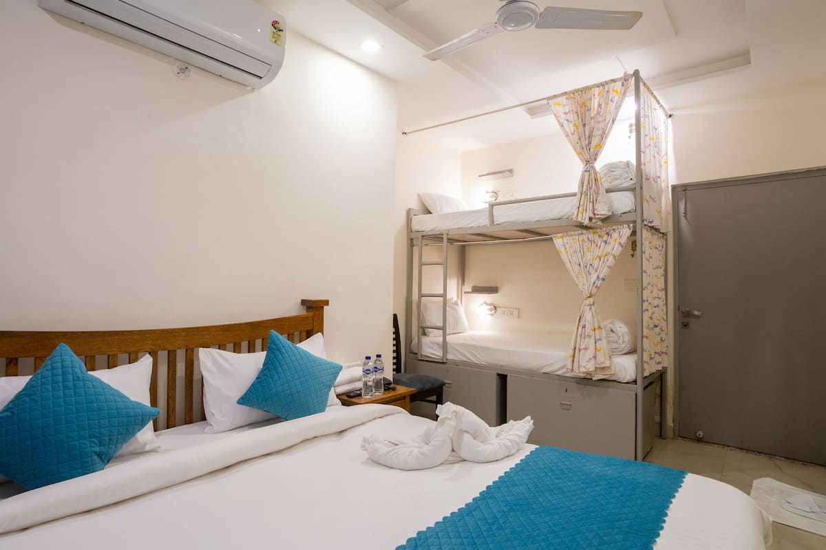 A hotel room with a single bed, a bunk bed, a wardrobe, a bedside table with a lamp, and an air conditioning unit.