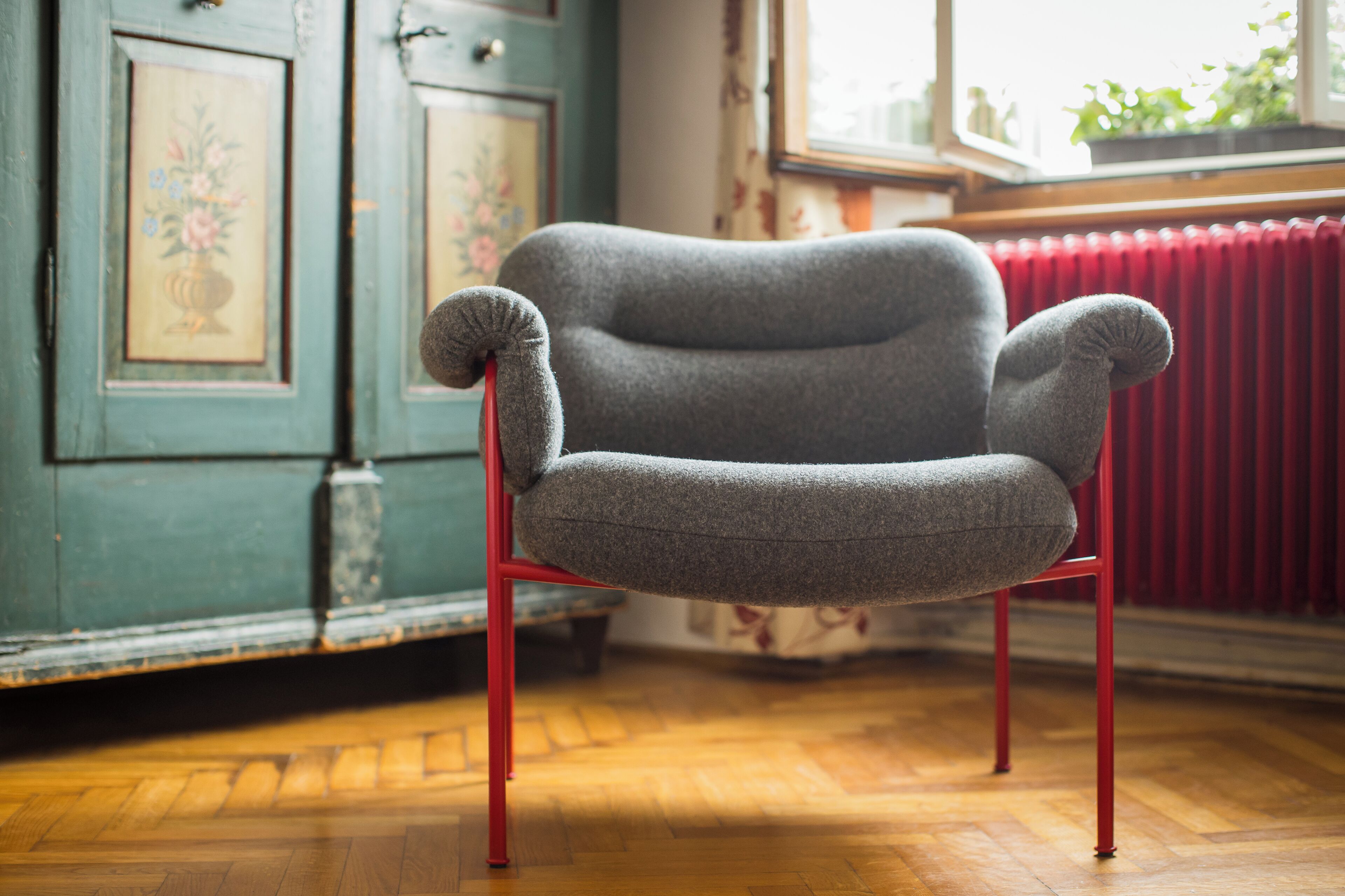 A grey armchair with red legs in a room with wooden flooring, a red radiator, and a window with plants.