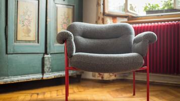 A grey armchair with red legs in a room with wooden flooring, a red radiator, and a window with plants.