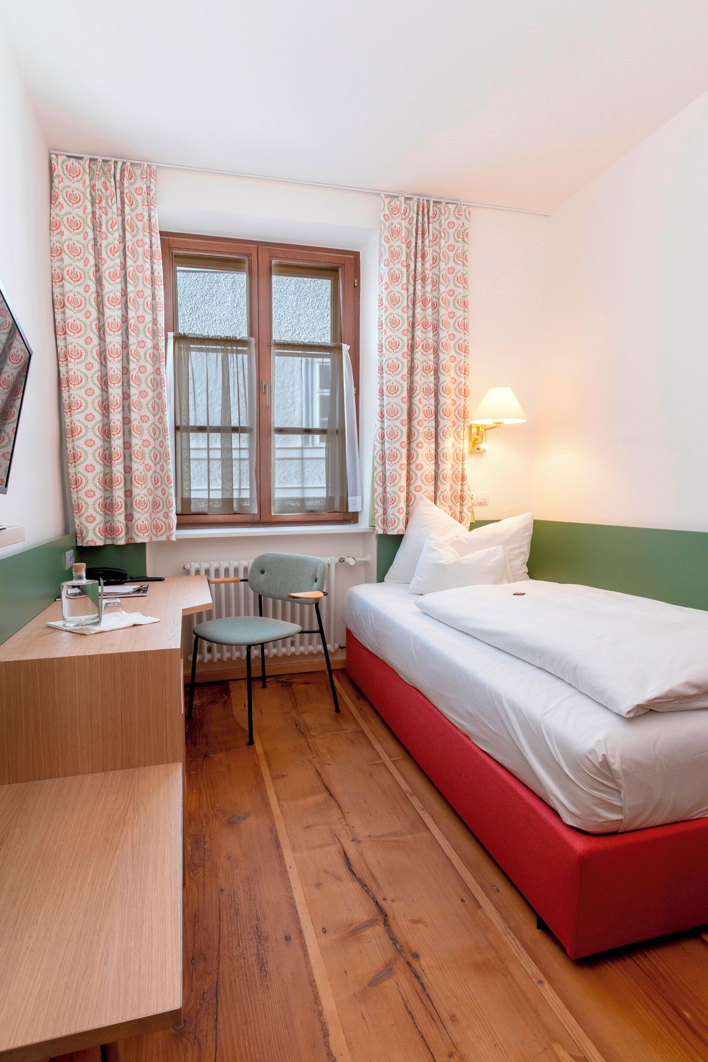 Una habitación con cama, escritorio, silla, ventana con cortinas y una lámpara de pared.