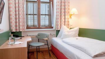 Una habitación con cama, escritorio, silla, ventana con cortinas y una lámpara de pared.
