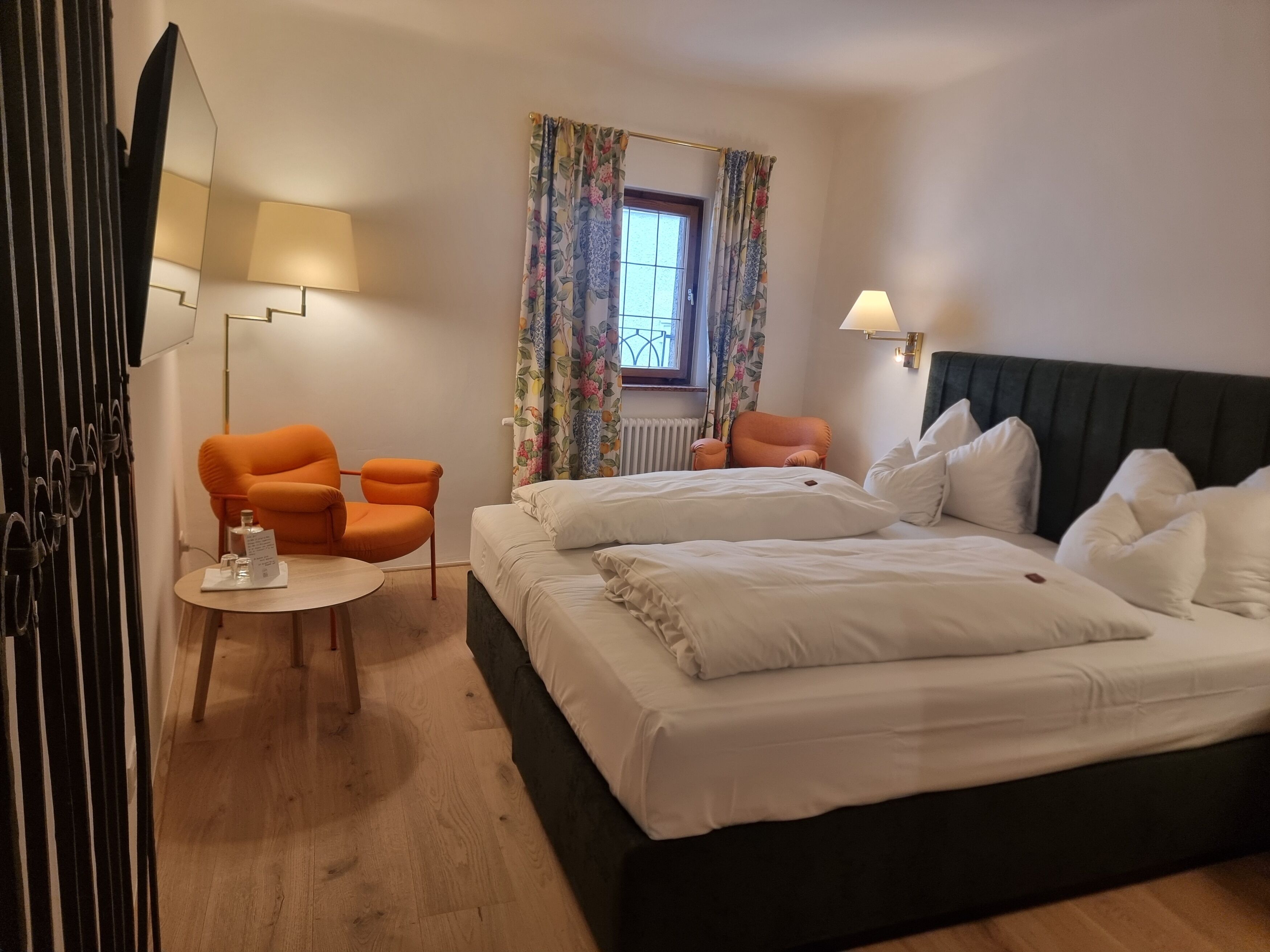 Habitación de hotel con cama, dos sillas, una mesita, lámpara y ventana con cortinas.