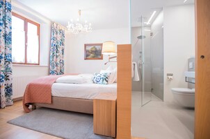 Un dormitorio moderno con cama, mesita de noche, candelabro, ventana con cortinas, un cuadro en la pared y un baño con inodoro y ducha.