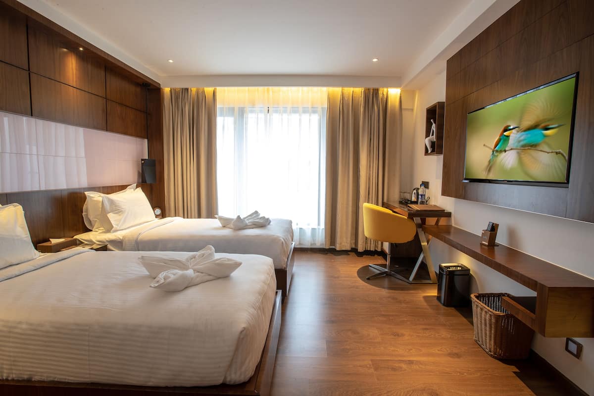 A hotel room with two beds, a desk, a television, and a large window with curtains.