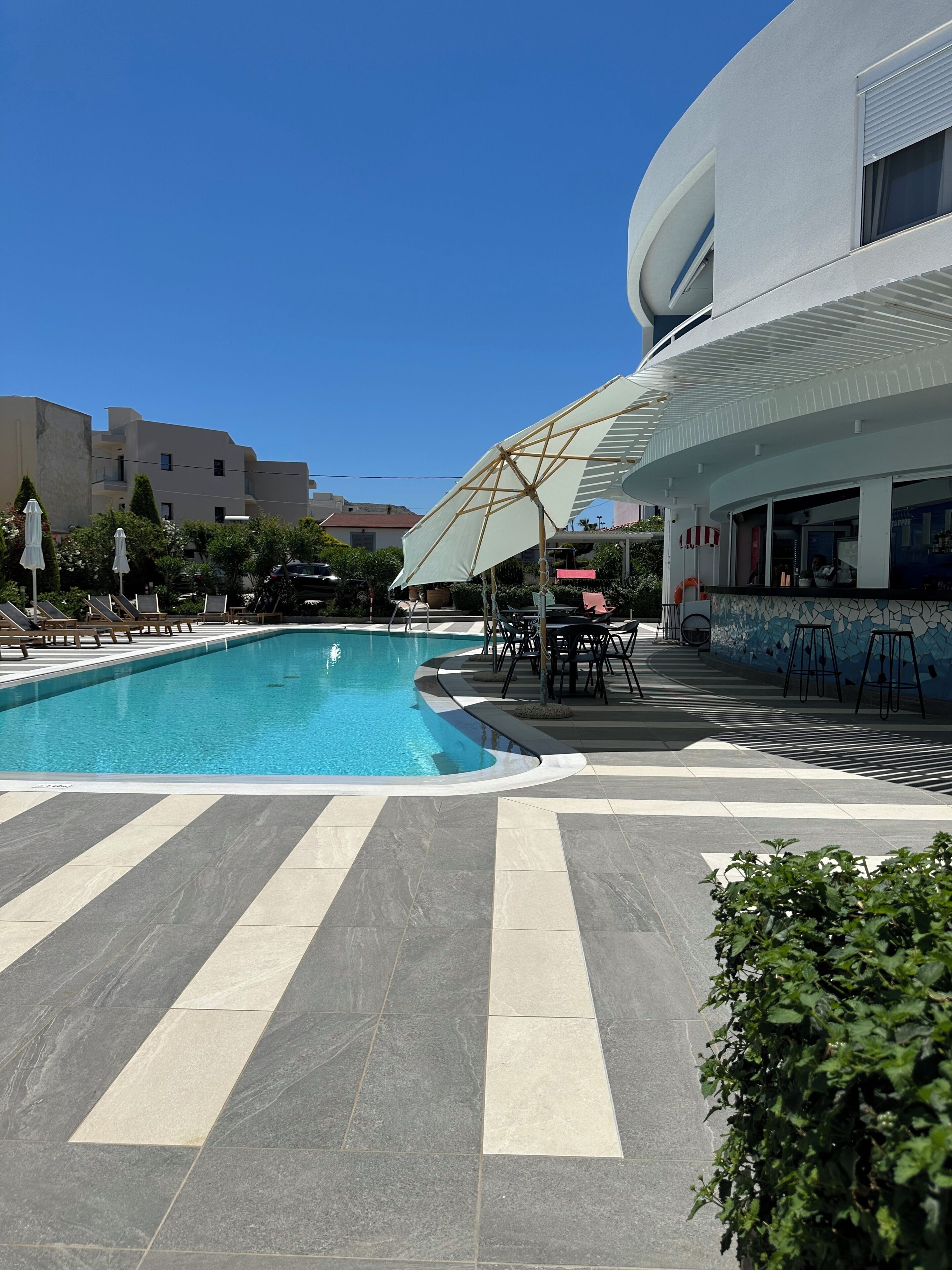 Una piscina con sillas de descanso y sombrillas, un espacio de estar cubierto y un edificio moderno con fachada de azulejos.