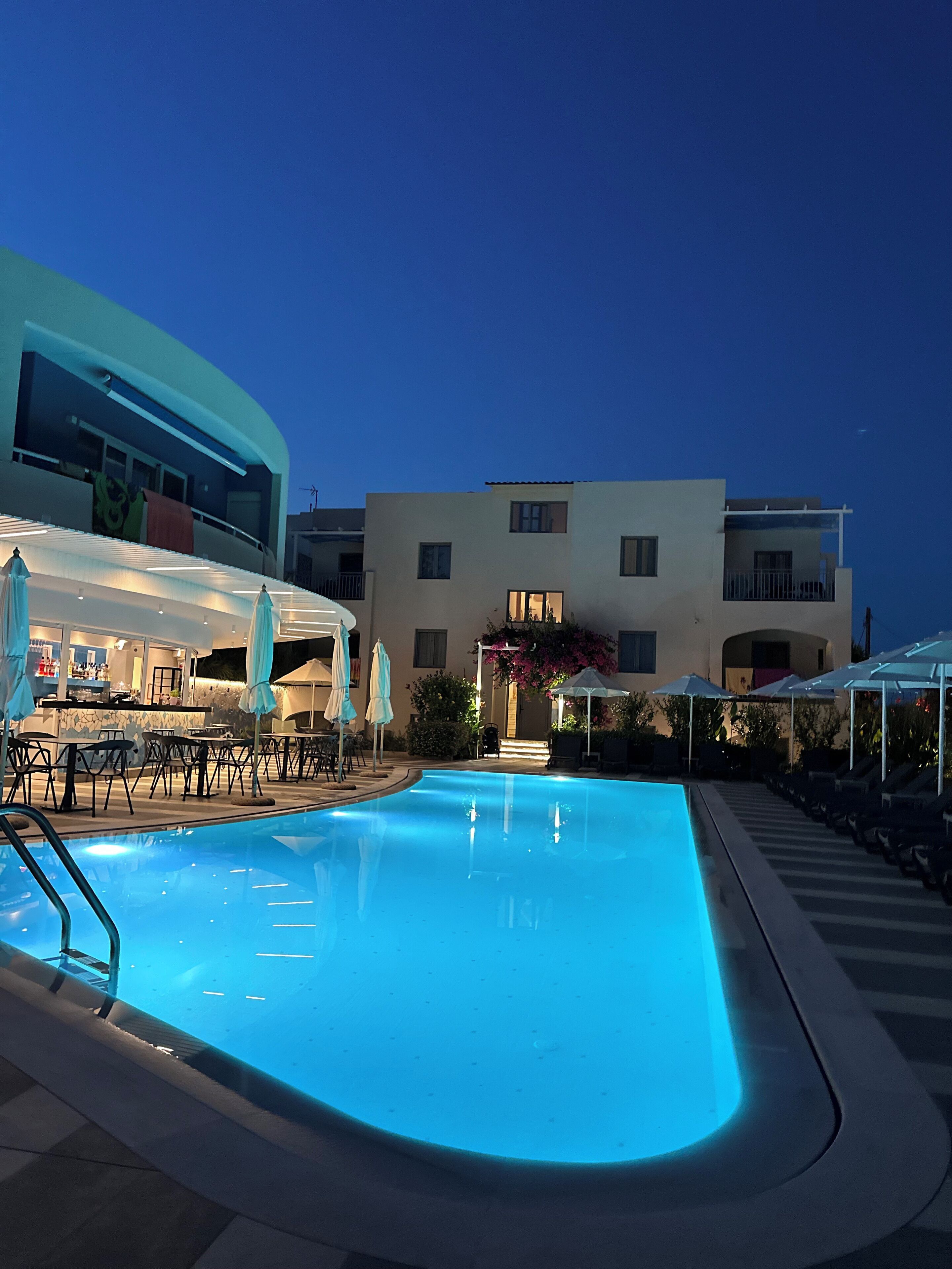 A swimming pool with lounge chairs and umbrellas, a bar area, and a building with multiple balconies in the background.