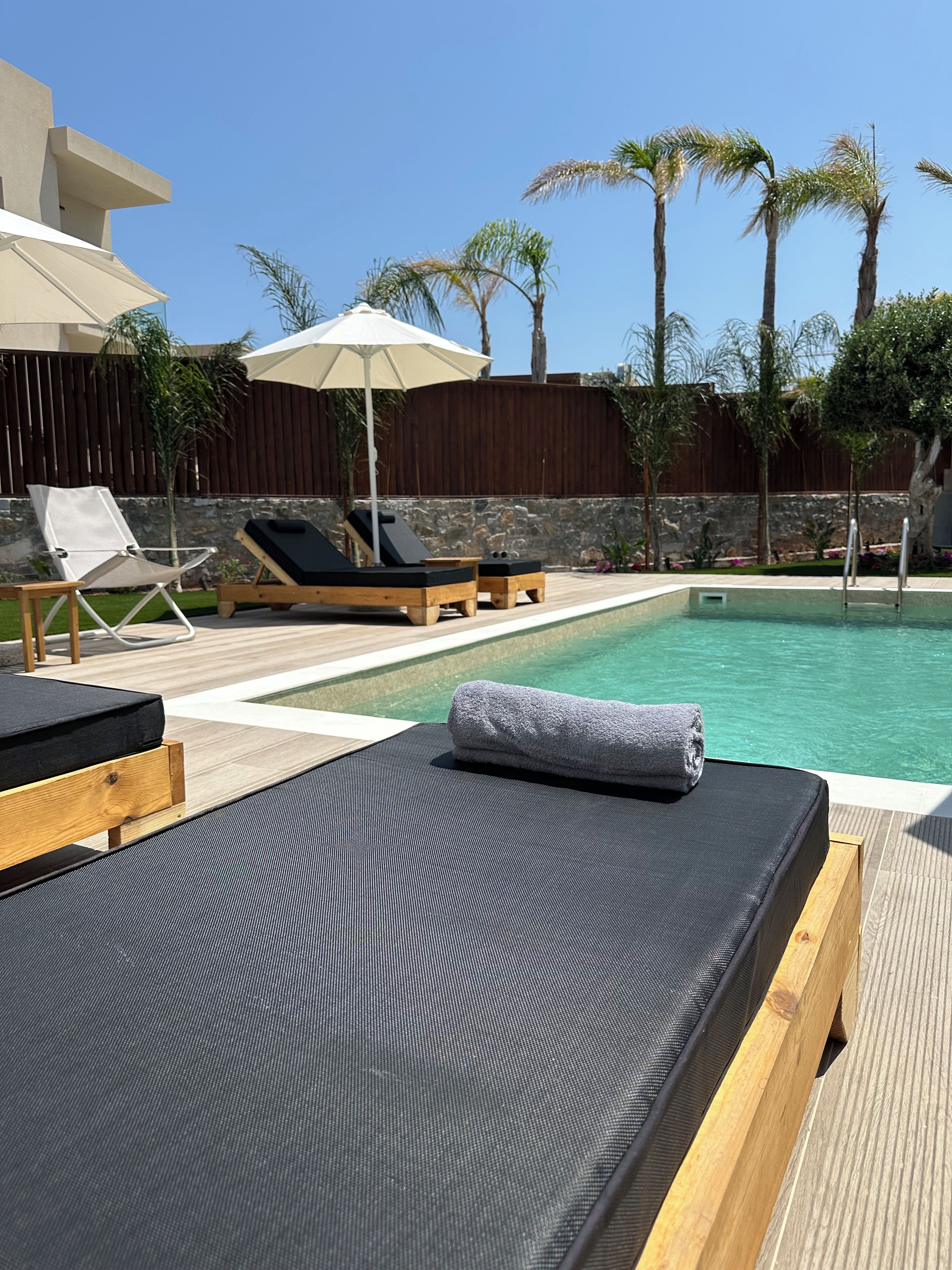 Un área junto a la piscina con sillones de descanso, una terraza de madera y un muro de piedra.