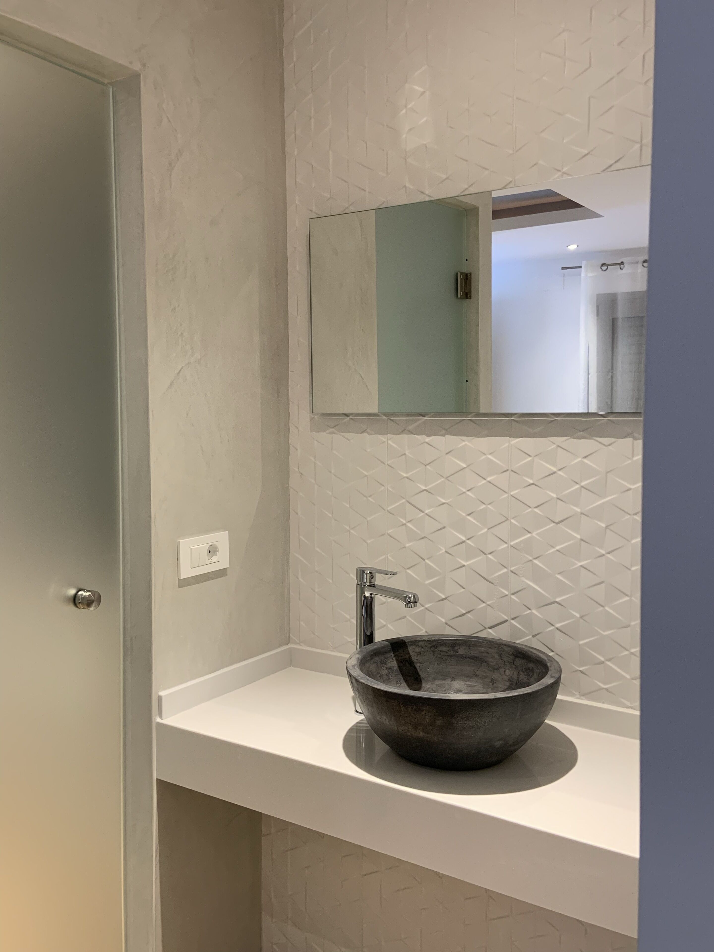 A bathroom with a white tiled wall, a large mirror, a round vessel sink, and a door.
