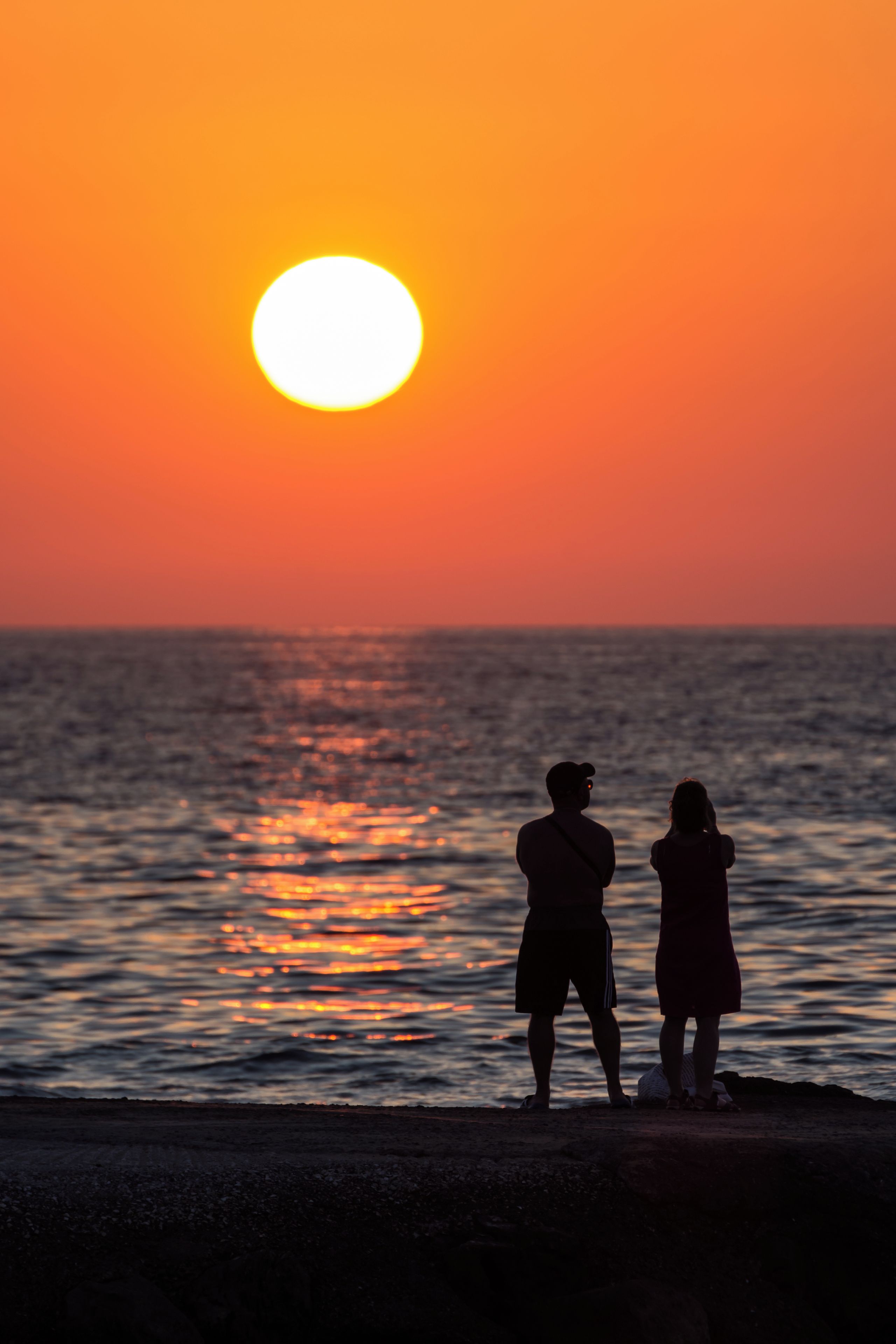 Two silhouettes of people standing by the sea during sunset.
