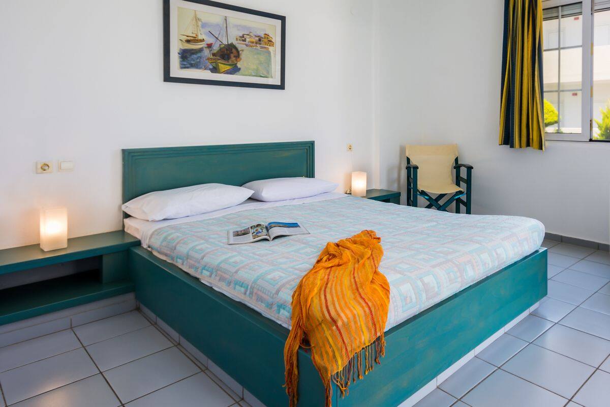 A bedroom with a teal bed, white pillows, a magazine on the bed, a yellow throw blanket, a chair, and a painting on the wall.